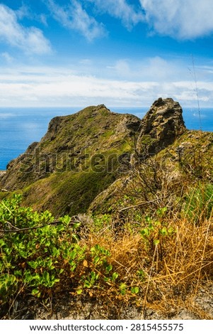 Dramatic volcanic peaks covered in vibrant green foliage meet the vast Atlantic Ocean under a bright blue sky with scattered clouds