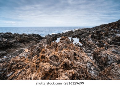 Dramatic volcanic rock formations line the shore of Tenerife, with calm tide pools reflecting the overcast sky above the vast ocean