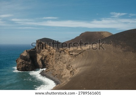 Dramatic volcanic sea cliffs with vivid layered geological strata rise above crashing Atlantic waves along a rugged coastline in the Azores, Portugal. Dark lava slopes and eroded sedimentary formations contrast sharply with the deep turquoise ocean under a partly cloudy blue sky, showcasing the raw volcanic origins of the archipelago. Ideal for travel, editorial, geotourism, and nature adventure content.