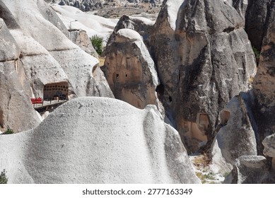 Dramatic volcanic tuff formations of Cappadocia's fairy chimneys near Ürgüp, showcasing troglodyte cave dwellings with carved entrances. Rustic wooden platform features vibrant red chairs, barrel, and lantern amid arid Anatolian landscape. Ideal for travel campaigns, geology books, adventure tourism ads highlighting Ancient Anatolia's Seljuk heritage in Osiana, Hagios Prokopios, Başköy.