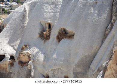 Dramatic volcanic tuff formations in Cappadocia's fairy chimneys feature ancient cave dwellings with rectangular window openings overgrown by wild grass and shrubs. Sunlit white rock faces tower over Ürgüp village backdrop with modern houses, cars, and construction amid arid Anatolian landscape. Evokes troglodyte heritage near Osiana, Hagios Prokopios, Başköy; ideal for Turkey travel campaigns, history books on Seljuk architecture, adventure ads alleviating urban escape cravings.