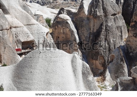 Dramatic volcanic tuff formations of Cappadocia's fairy chimneys near Ürgüp, showcasing troglodyte cave dwellings with carved entrances. Rustic wooden platform features vibrant red chairs, barrel, and lantern amid arid Anatolian landscape. Ideal for travel campaigns, geology books, adventure tourism ads highlighting Ancient Anatolia's Seljuk heritage in Osiana, Hagios Prokopios, Başköy.