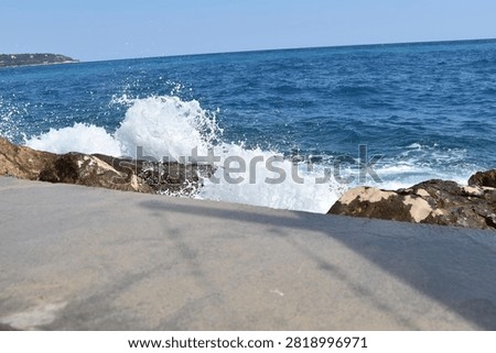 A dramatic wave explodes into a tall arc of white spray as it strikes dark, rugged rocks along the shoreline. Beyond the splash, the sea deepens into rich navy tones under a hazy sky, remaining calm in contrast to the sudden burst of energy near the rocks. Captured from a paved coastal path, the image freezes a fleeting moment of powerful motion against the stillness of the open water beyond, full of texture and dynamic contrast. Great for travel, nature, or action-oriented coastal content.
