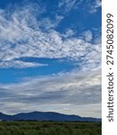 Dramatic white clouds across blue sky above mountain range and green grass field. Scenic natural landscape with wide copy space. Peaceful outdoor countryside environment in daylight.