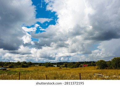 Dramatic white cumulus clouds drift across a vibrant blue sky above a sprawling golden meadow with lush greenery and distant village houses under a bright summer sun