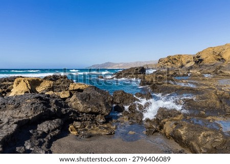 A dramatic wide-angle perspective showcases the untamed beauty of La Pared beach with dark sand and scattered golden boulders. Crashing turquoise waves roll into the shore under a massive, clear blue sky, offering excellent copy space for travel concepts.