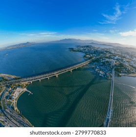 A dramatic wide-angle shot looking up at the massive concrete pillars and structure of a modern highway bridge stretching over the rippling ocean water, with a distant city coastline glowing under the bright afternoon sun.