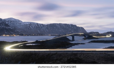 Dramatic wide-angle view of the Atlantic coast road in Lofoten with glowing light streaks from vehicles at dusk.
