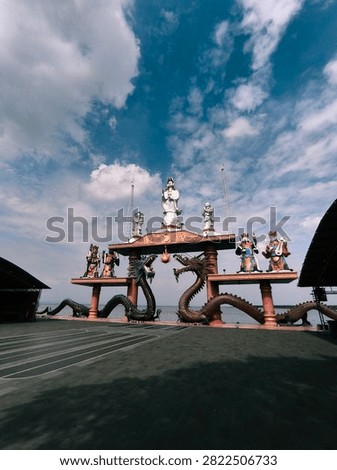 A dramatic wide-angle view of a traditional Chinese temple gate featuring majestic dragon sculptures and towering statues of deities against a vibrant blue sky filled with dynamic clouds. Located by the waterfront, this iconic cultural landmark showcases intricate architecture, colorful religious symbolism, and impressive craftsmanship.