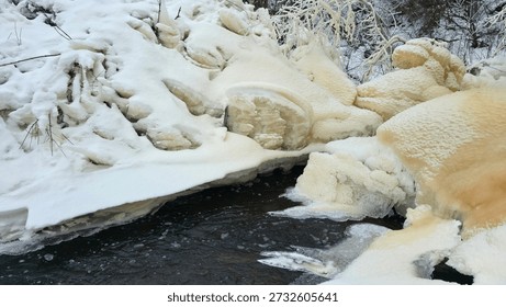 Dramatic winter ice formations along frozen stream in Norway. Snow-covered rocks and sculptural ice chunks with golden hues beside dark flowing water. Natural winter landscape showing contrast between frozen ice structures and open water creating unique patterns and textures in Nordic nature.