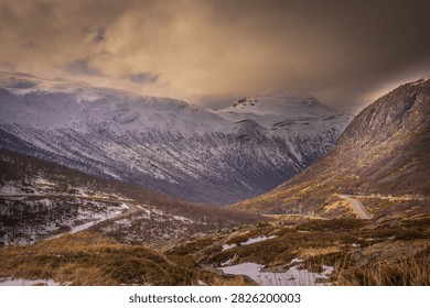 Dramatic winter landscape along E134 at Haukelifjell in Norway, with a winding mountain road leading through snow-covered peaks and rugged alpine terrain. Warm evening light illuminates the mountains beneath dark storm clouds, creating a striking Scandinavian travel and landscape scene.