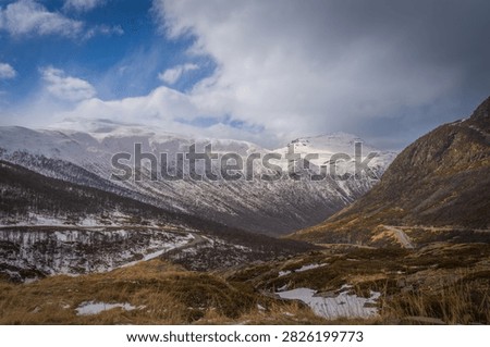 Dramatic winter landscape along E134 at Haukelifjell in Norway, with a winding mountain road leading through snow-covered peaks and rugged alpine terrain. Warm evening light illuminates the mountains beneath dark storm clouds, creating a striking Scandinavian travel and landscape scene.