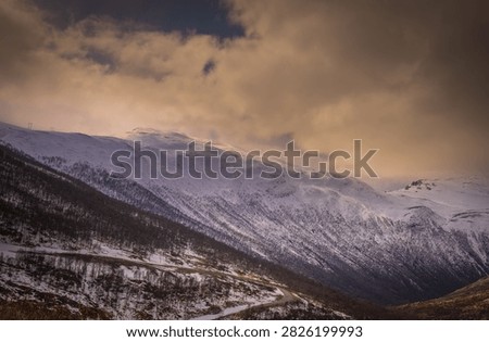 Dramatic winter landscape along E134 at Haukelifjell in Norway, with a winding mountain road leading through snow-covered peaks and rugged alpine terrain. Warm evening light illuminates the mountains beneath dark storm clouds, creating a striking Scandinavian travel and landscape scene.