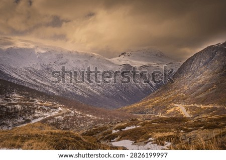 Dramatic winter landscape along E134 at Haukelifjell in Norway, with a winding mountain road leading through snow-covered peaks and rugged alpine terrain. Warm evening light illuminates the mountains beneath dark storm clouds, creating a striking Scandinavian travel and landscape scene.