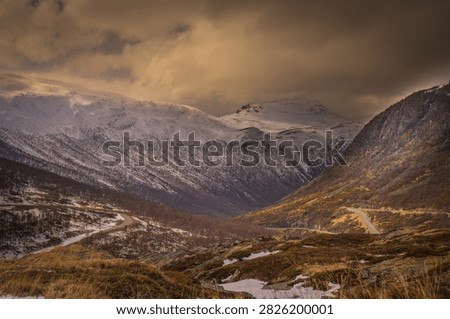 Dramatic winter landscape along E134 at Haukelifjell in Norway, with a winding mountain road leading through snow-covered peaks and rugged alpine terrain. Warm evening light illuminates the mountains beneath dark storm clouds, creating a striking Scandinavian travel and landscape scene.