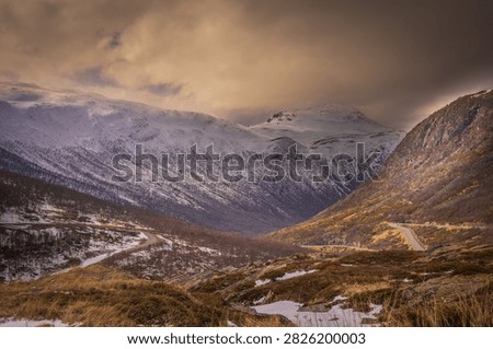 Dramatic winter landscape along E134 at Haukelifjell in Norway, with a winding mountain road leading through snow-covered peaks and rugged alpine terrain. Warm evening light illuminates the mountains beneath dark storm clouds, creating a striking Scandinavian travel and landscape scene.
