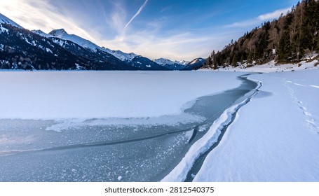 A dramatic winter landscape featuring a partially frozen alpine lake with a prominent crack in the ice, surrounded by snow-covered mountains and pine forests under a clear sky.