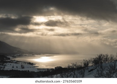 Dramatic winter light breaks through dark clouds over Malangen fjord near Tromso, Norway. Sun rays reflect on calm water, creating cinematic Arctic landscape with moody atmosphere.