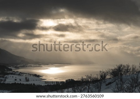 Dramatic winter light breaks through dark clouds over Malangen fjord near Tromso, Norway. Sun rays reflect on calm water, creating cinematic Arctic landscape with moody atmosphere.