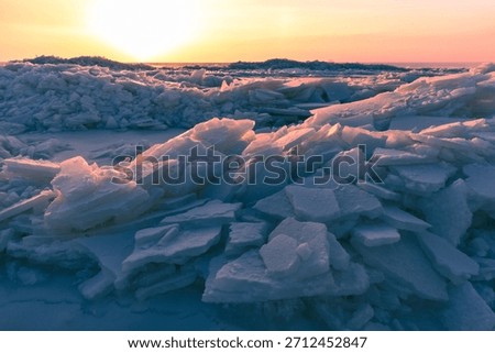 A dramatic winter scene shows broken ice chunks piled along a frozen sea, bathed in warm sunset colors. The blue shadows contrast with orange light, creating a tranquil, chilly, and awe-inspiring mood