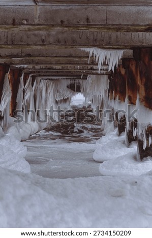A dramatic winter scene under a pier: icicles hang from concrete pillars, forming a natural ice tunnel over flowing water. The cold, moody atmosphere captures the raw beauty of frozen nature.