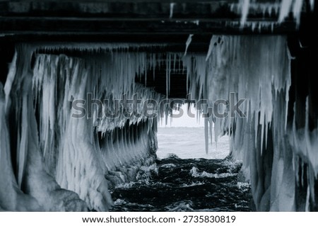 A dramatic winter scene under a pier: icicles hang from concrete pillars, forming a natural ice tunnel over flowing water. The cold, moody atmosphere captures the raw beauty of frozen nature.