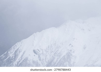 A dramatic winter scene of a white alpine mountain slope covered in snow, sharply contrasting against a dark gray stormy sky with fog and mist.