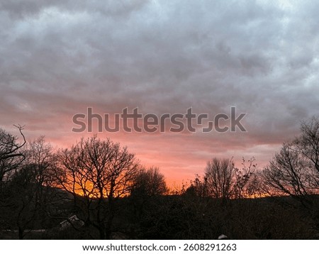 
A dramatic winter sunset with fiery orange and pink hues glowing beneath a blanket of grey clouds, silhouetting bare trees in a peaceful countryside landscape.
