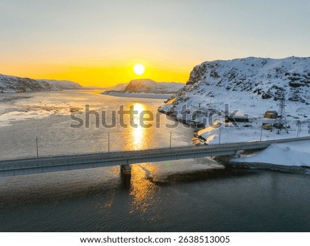 Dramatic winter sunset over the Teriberka river, with a bridge connecting snow-covered banks and distant mountains, Russia