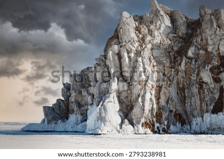 Dramatic winter view of Shaman Rock on Olkhon Island at Lake Baikal, Russia, with frozen shoreline, rugged cliffs and dark storm clouds over the ice