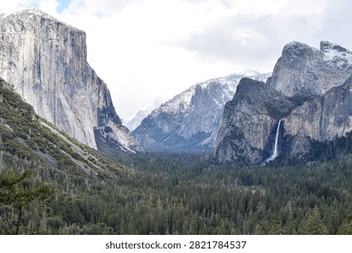 A dramatic winter view of Yosemite Valley from Tunnel View in Yosemite National Park, California, featuring El Capitan, Bridalveil Fall and snow-covered granite peaks beneath storm clouds breaking to reveal blue sky.