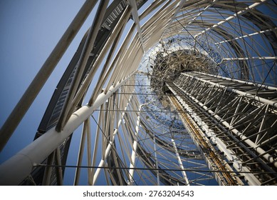 A dramatic worm's-eye view looking up through the intricate white steel framework of a modern tower. The complex network of pipes and cables creates a rhythmic geometric pattern, leading the eye toward a glass-domed top under a bright, clear sky.