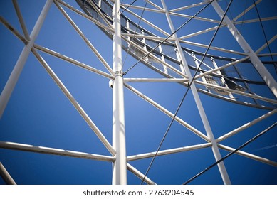 A dramatic worm's-eye view looking up through the intricate white steel framework of a modern tower. The complex network of pipes and cables creates a rhythmic geometric pattern, leading the eye toward a glass-domed top under a bright, clear sky.