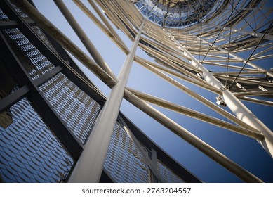 A dramatic worm's-eye view looking up through the intricate white steel framework of a modern tower. The complex network of pipes and cables creates a rhythmic geometric pattern, leading the eye toward a glass-domed top under a bright, clear sky.