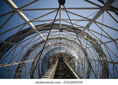 A dramatic worm's-eye view looking up through the intricate white steel framework of a modern tower. The complex network of pipes and cables creates a rhythmic geometric pattern, leading the eye toward a glass-domed top under a bright, clear sky.