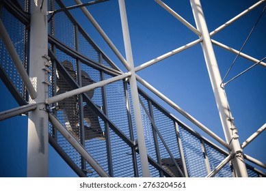 A dramatic worm's-eye view looking up through the intricate white steel framework of a modern tower. The complex network of pipes and cables creates a rhythmic geometric pattern, leading the eye toward a glass-domed top under a bright, clear sky.