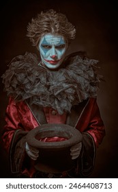 A dramatic young man - actor with clown, joker makeup takes off his hat to beg and looks at the camera with a heavy, embittered look. Black background. Drama, tragedy. Theater and cinema.