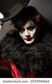 A dramatic young man - an actor with clown, joker makeup looking to the camera with tired eyes and a smile. Black background. Drama, tragedy. Performance in the theater.