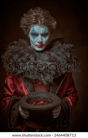 A dramatic young man - actor with clown, joker makeup takes off his hat to beg and looks at the camera with a heavy, embittered look. Black background. Drama, tragedy. Theater and cinema.