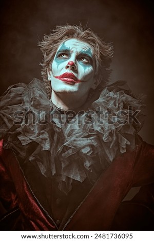 A dramatic young man - an actor with clown, joker makeup looking up to the light with tired eyes. Black background. Drama, tragedy. Performance in the theater.