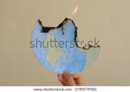 Drawing of the Earth, on fire. The planet is burning, literally from Global Warming, figuratively from hate, greed, racism, terrorism, and wars. Cut out is held up by a hand, against a blank wall.