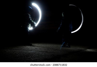 Drawing With Light At Night With Long Exposure. Spiral Circles And Wavy Lines Using Pyrotechnic Sparklers Led Lights. White Color Effect Hand Movements Of Children, Lightsabers, Fencing