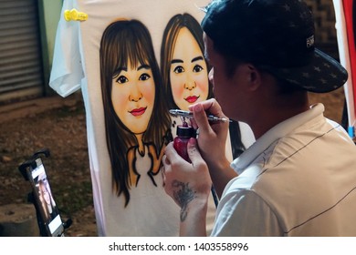 Drawing On Clothes. Man Draws Portrait Girls On A White T-shirt. Street Artist Asian At Work. January 16, 2019. Vang Vieng, Laos.