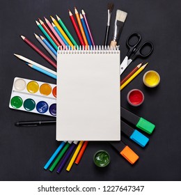 
Drawing supplies, empty drawing pad, crayons, paintbrushes, watercolor paints  on blackboard. 