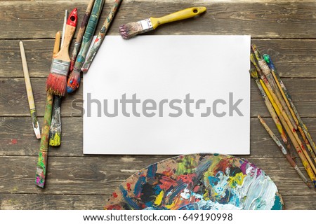 Drawing tools in art studio. Top view photo of paintbrushes next to palette with oil paints brushstrokes mixture and mockup of paper. Artist old wooden table with accessories. Creative concept