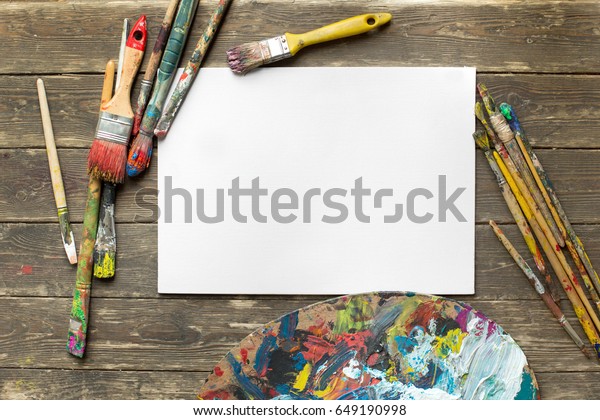 Drawing tools in art studio. Top view photo of\
paintbrushes next to palette with oil paints brushstrokes mixture\
and mockup of paper. Artist old wooden table with accessories.\
Creative concept