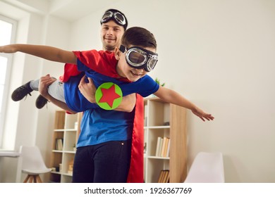 Dream Big. Loving Father Helps His Son Fly Like A Superhero. Boy With Pilot Glasses Play Fly With His Dad At Home. Cheerful Family In Red Cloaks And Pilot Glasses Are Having Fun Together.