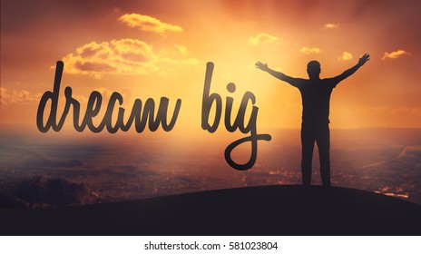 "Dream big" text on sunset background. Silhouette of man with raised hands on top of a mountain enjoying the view.