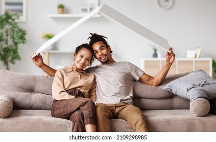 Dream Home. Portrait Of Young Happy African American Married Couple Holding White House Roof Overhead While Sitting On Sofa In New Flat, Smiling Black Family Man And Woman Getting First Apartment