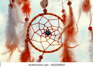 Dreamcatcher with orange feathers, swaying with light wind against background of evening city buildings, blue sky at sunset summer. Boho style, Indian style. Relax. Meditation, emotions